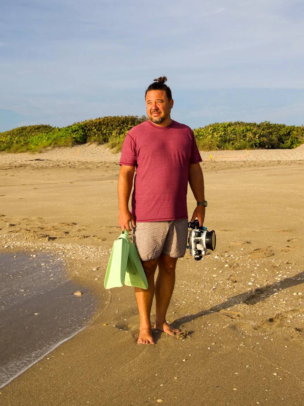 Brian Robinson standing barefoot at the waterline on a Florida beach at golden hour, swim fins in one hand and an underwater camera housing in the other.