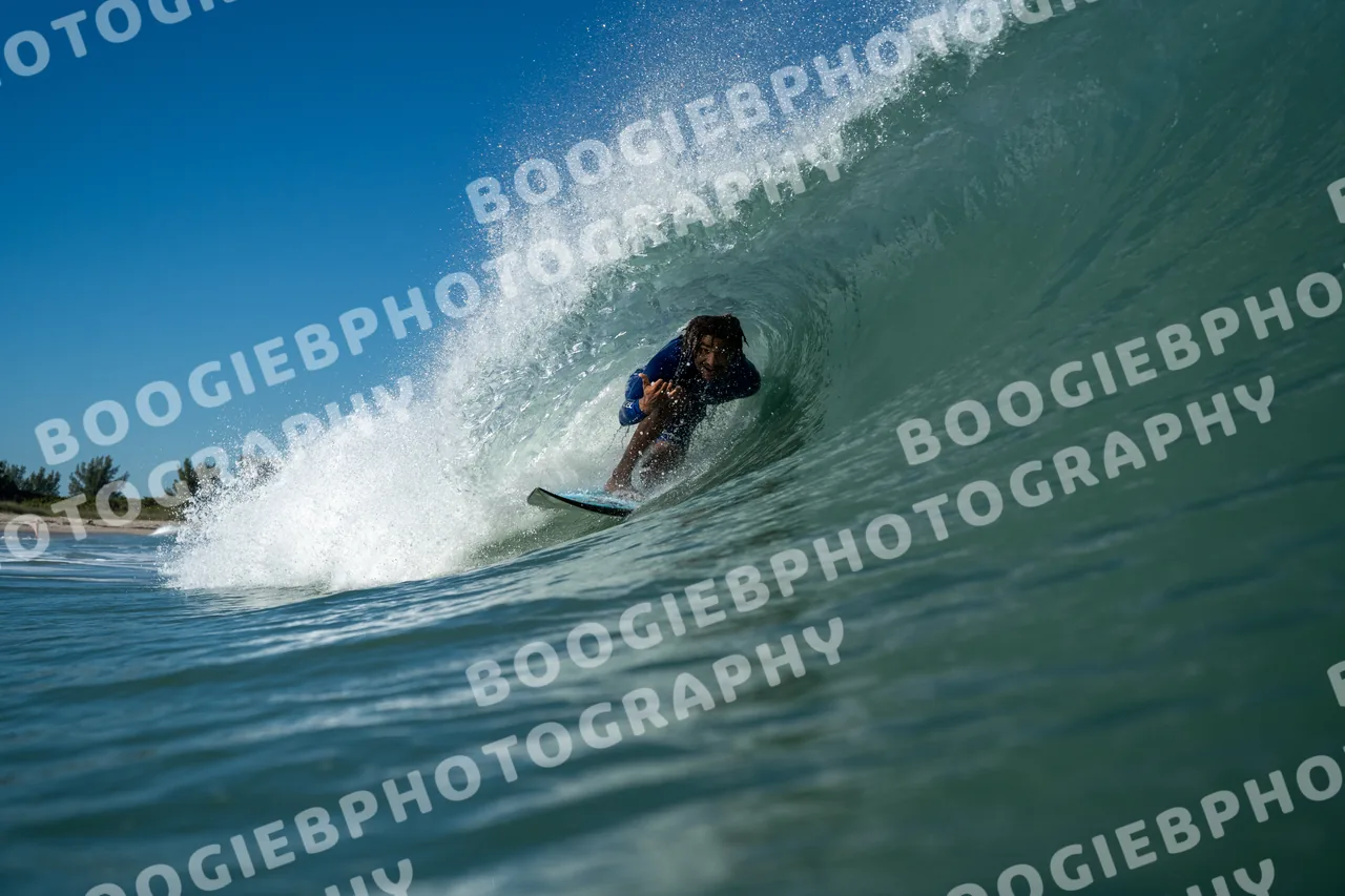 Surfer crouched inside a clean blue-green barrel, spray fanning off the lip, a low treeline on the horizon.