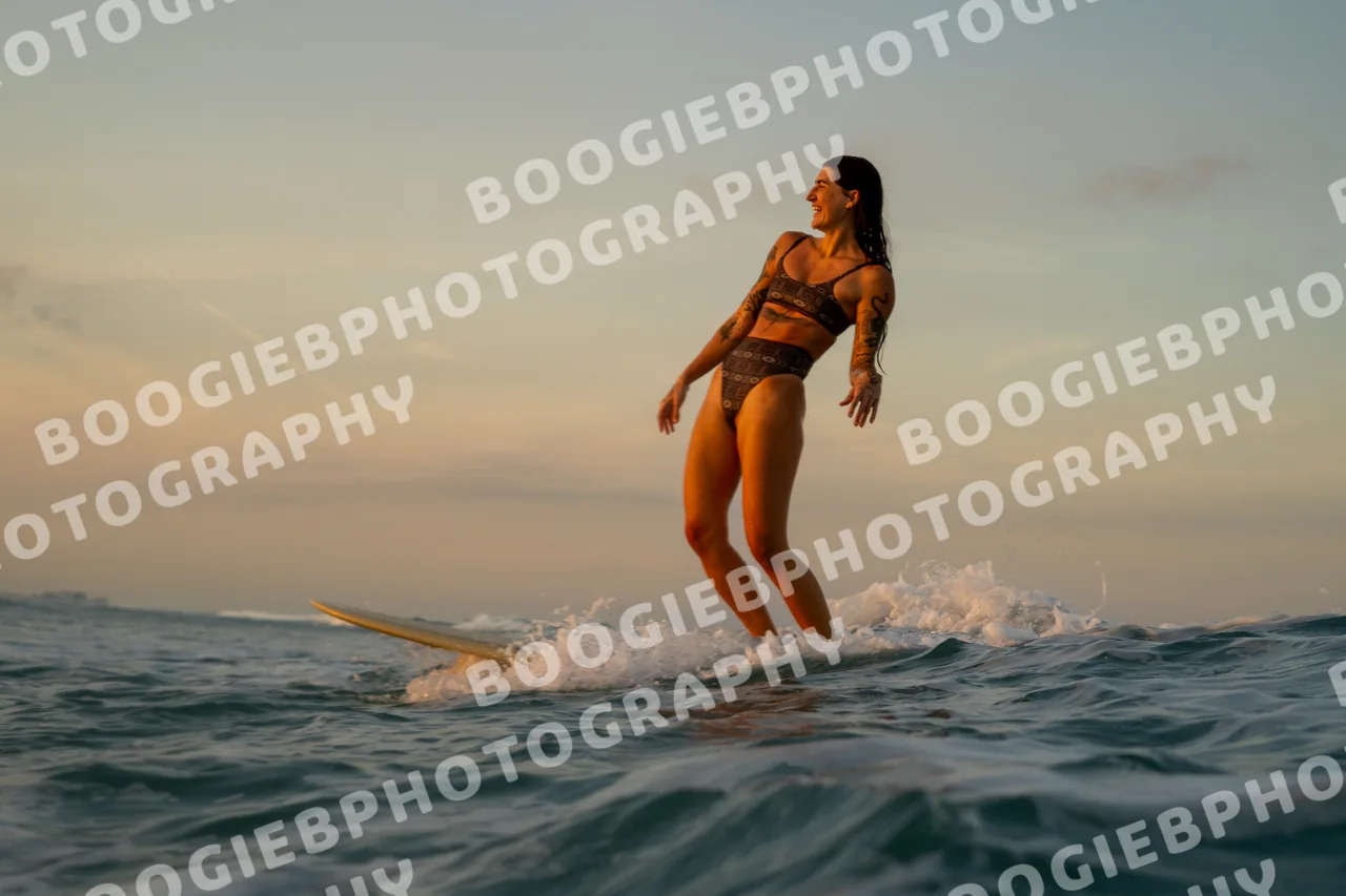 Longboarder standing tall on a pale board at sunset, stepping toward the nose, pastel sky behind.