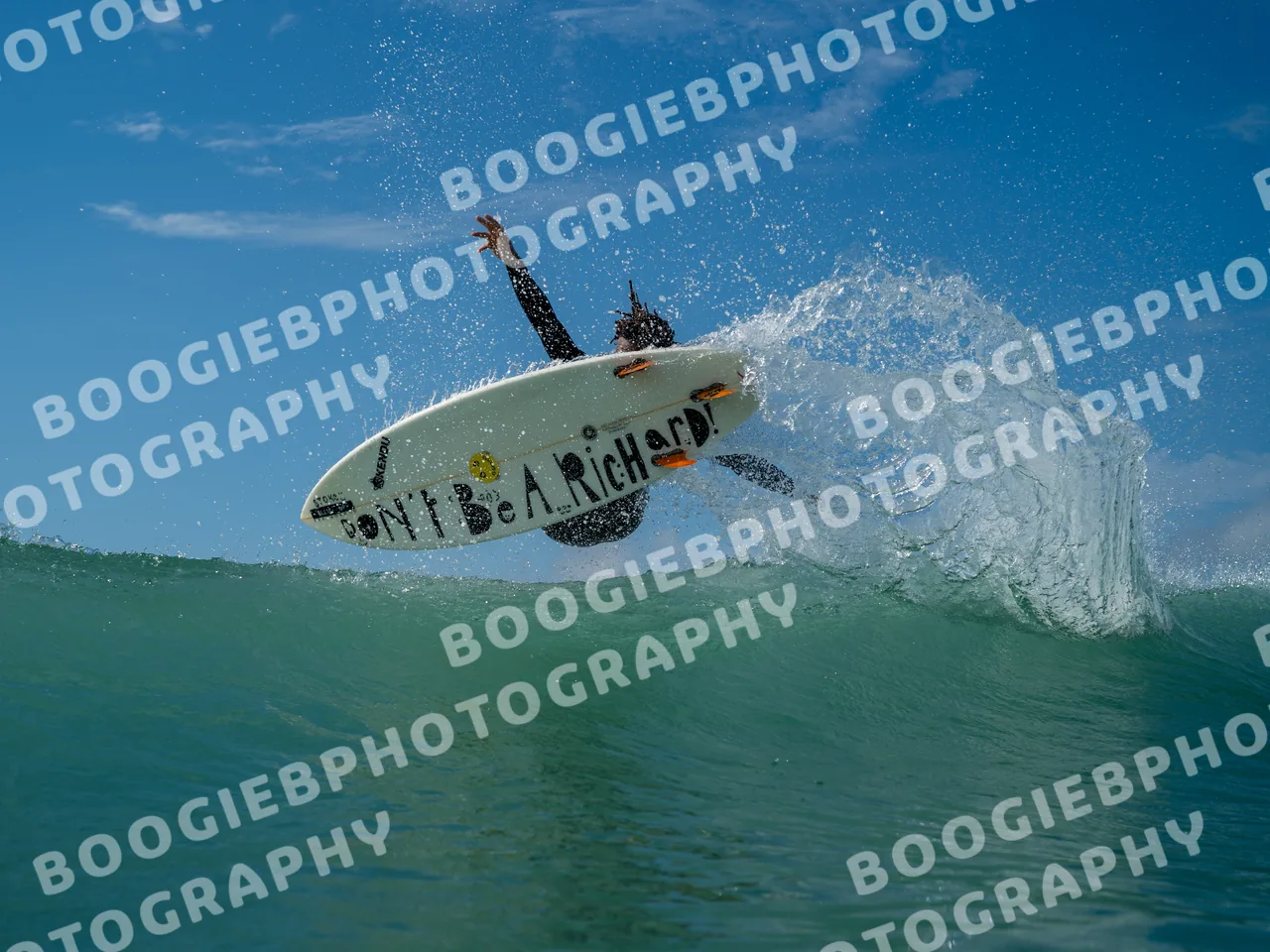 Surfer launching off the lip with one arm raised, the board turned to show a hand-lettered deck reading "DON'T BE A RICHARD!" against blue sky.