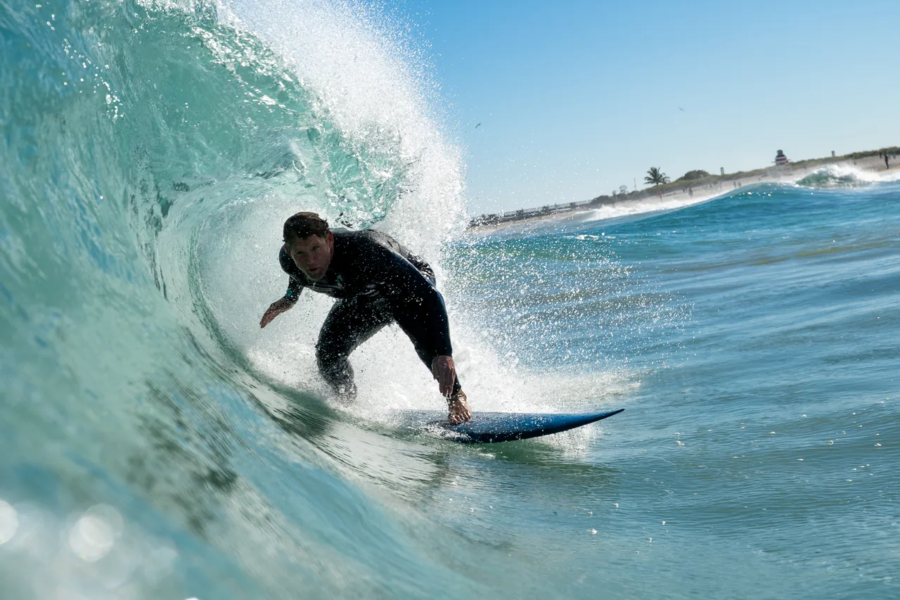 Surfer in a black wetsuit driving out of a hollow section, hand dragging in the face, beach and palms behind.