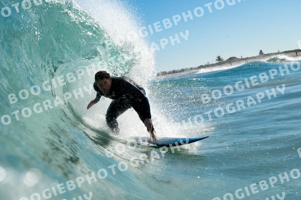 Surfer in a black wetsuit driving out of a hollow section, hand dragging in the face, beach and palms behind.