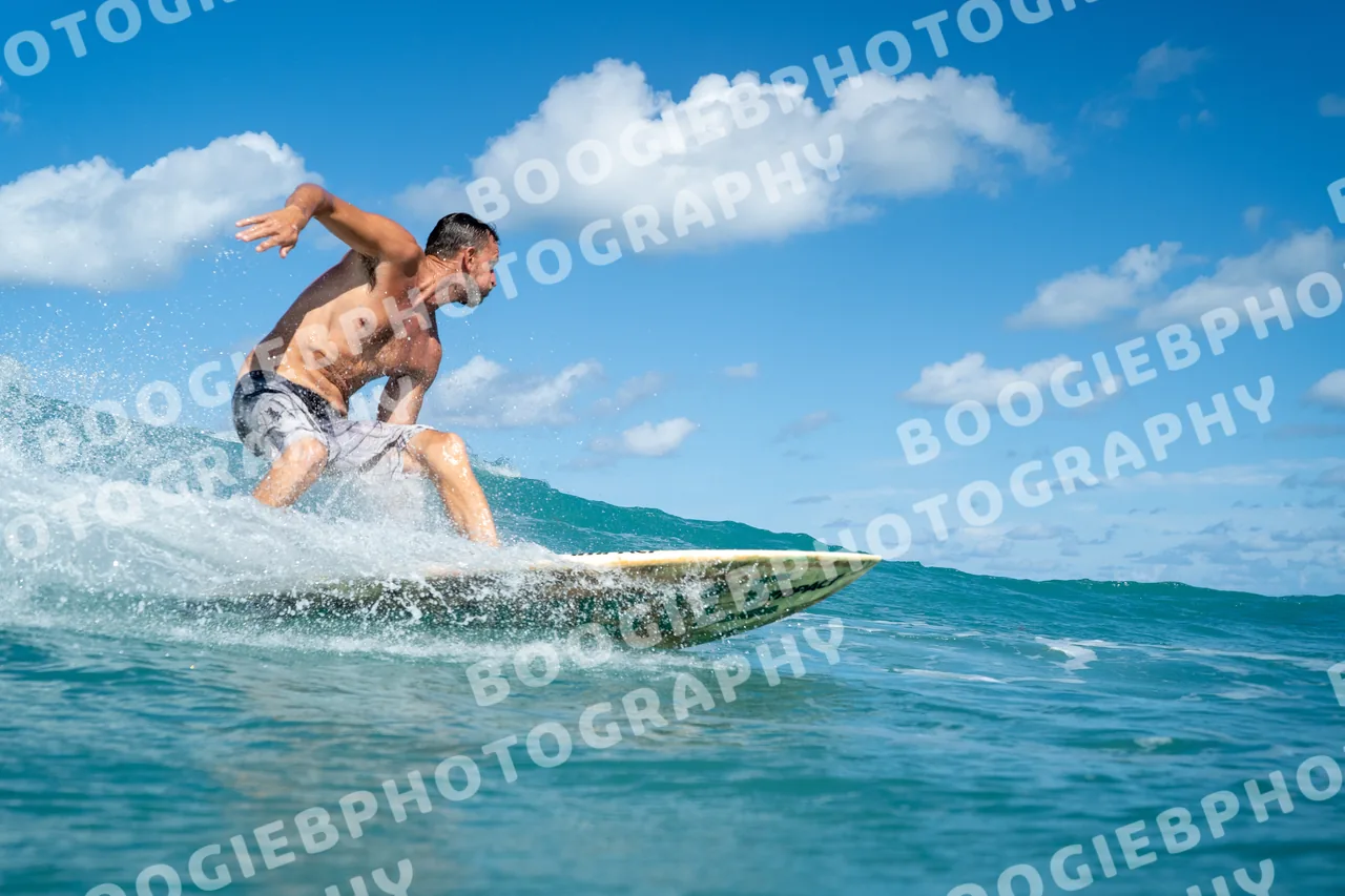 Surfer in board shorts crouched low on a longboard across a small bright wave, trade-wind clouds behind.