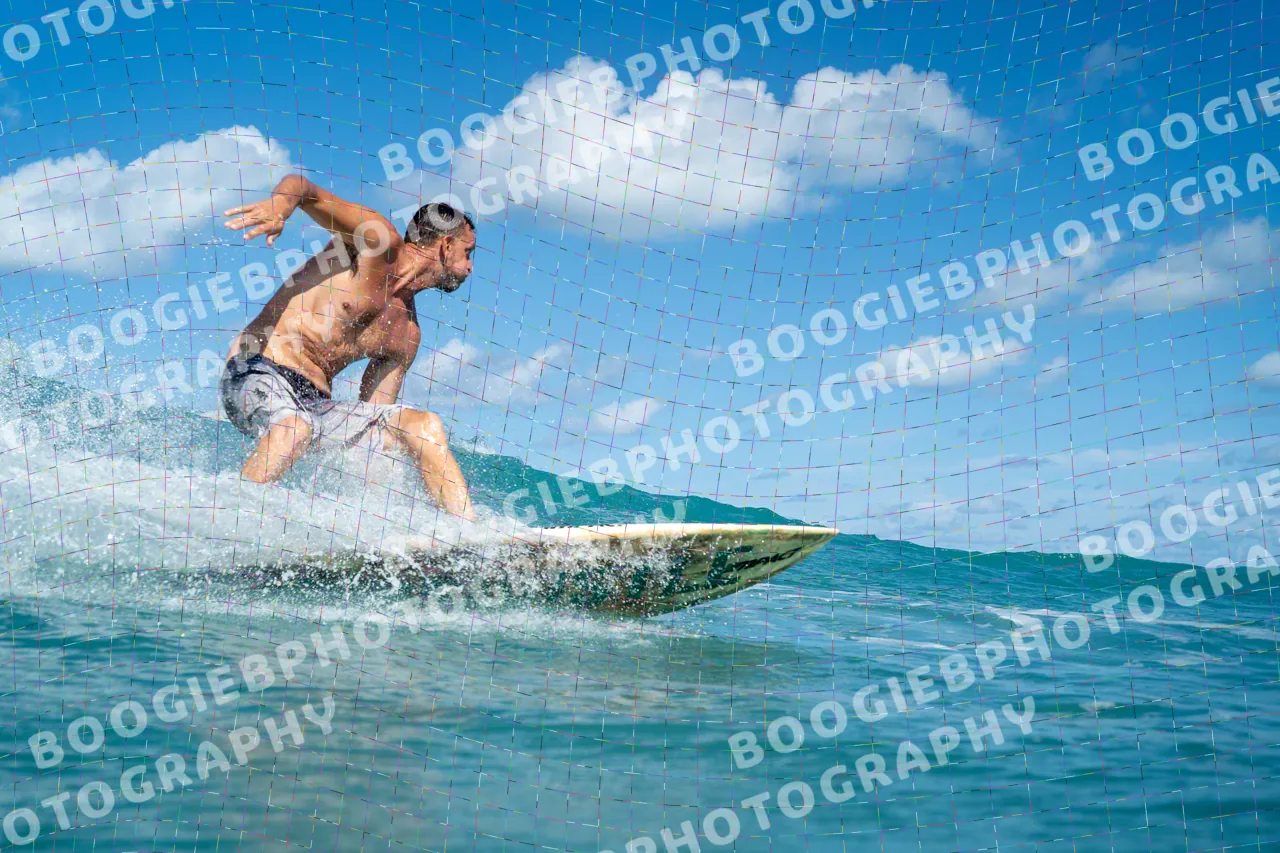 Surfer in board shorts crouched low on a longboard across a small bright wave, trade-wind clouds behind.