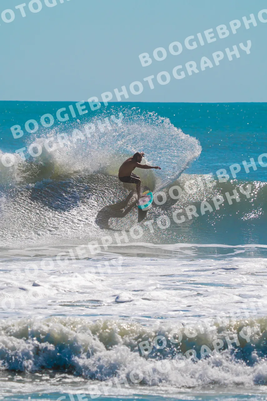 Surfer snapping off the top of a small teal wave, shot from the beach across lines of whitewater.