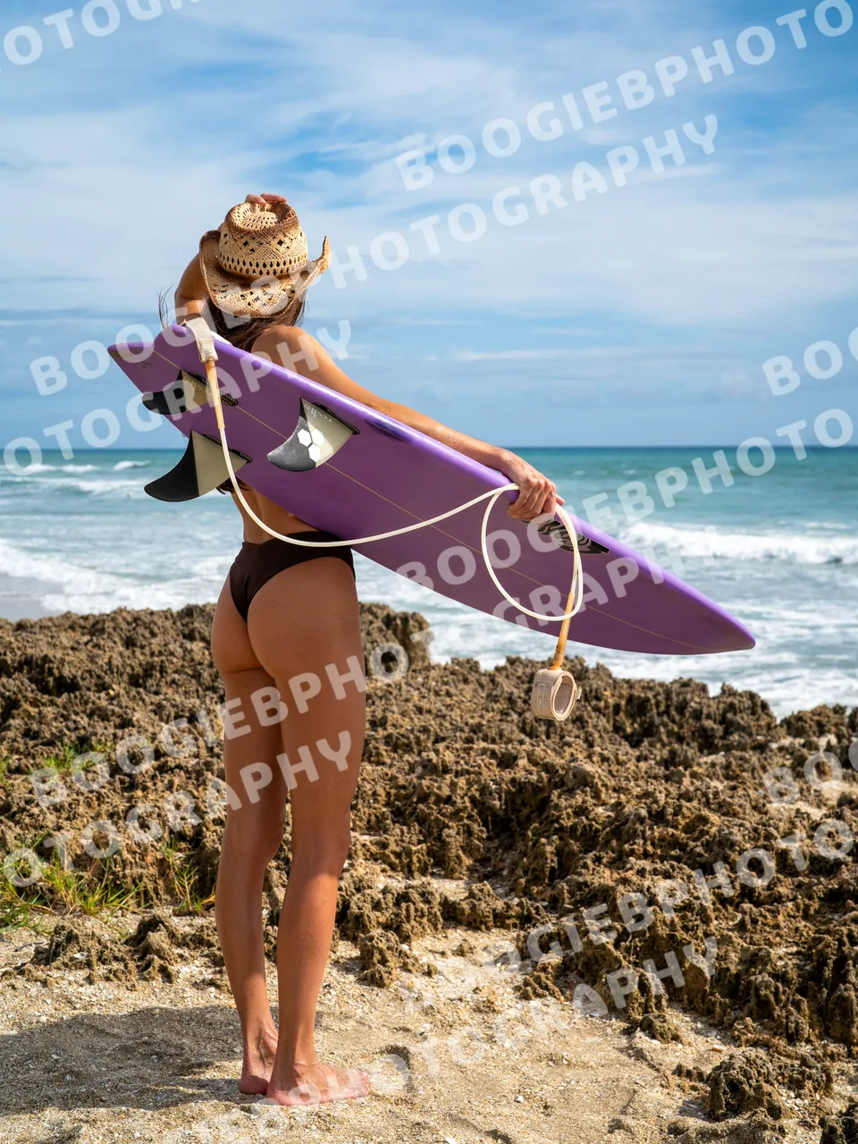 Model seen from behind on a rock shelf, the purple surfboard carried under one arm, looking out at breaking surf.