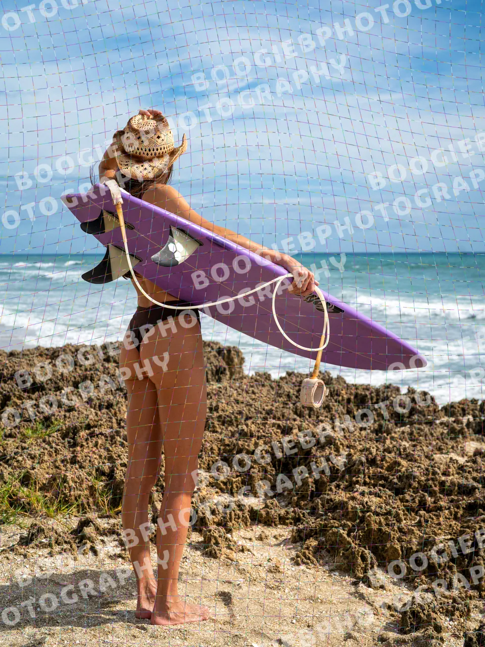 Model seen from behind on a rock shelf, the purple surfboard carried under one arm, looking out at breaking surf.